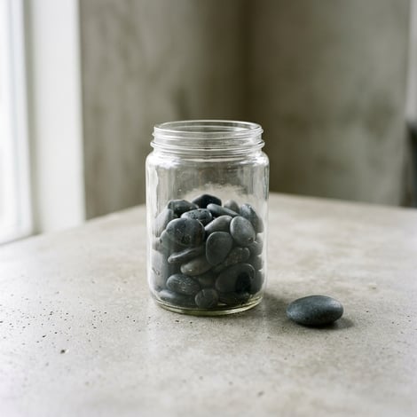 Square compHelp Others in B2B Networking: Give Before You Receiveosition: A close-up of a simple, clear glass jar half-filled with smooth, dark pebbles, placed on a neutral-toned table. A single pebble rests outside the jar, as if offered. Diffused overh